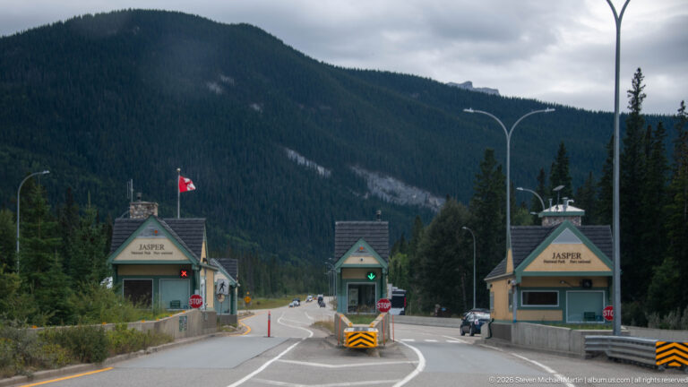 Jasper entrance booths spanning a divided roadway with stop signs, lane signals, Canadian flag, and forested mountains behind.