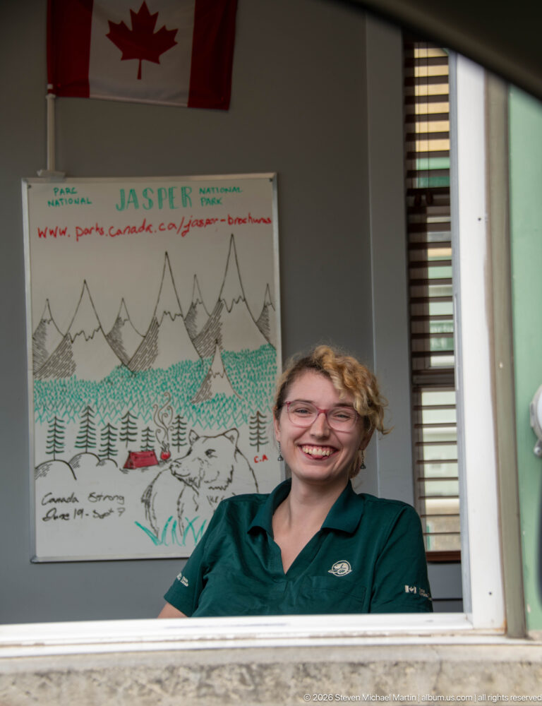 Smiling uniformed staff member at a service window with a Canadian flag and hand-drawn Jasper poster behind.