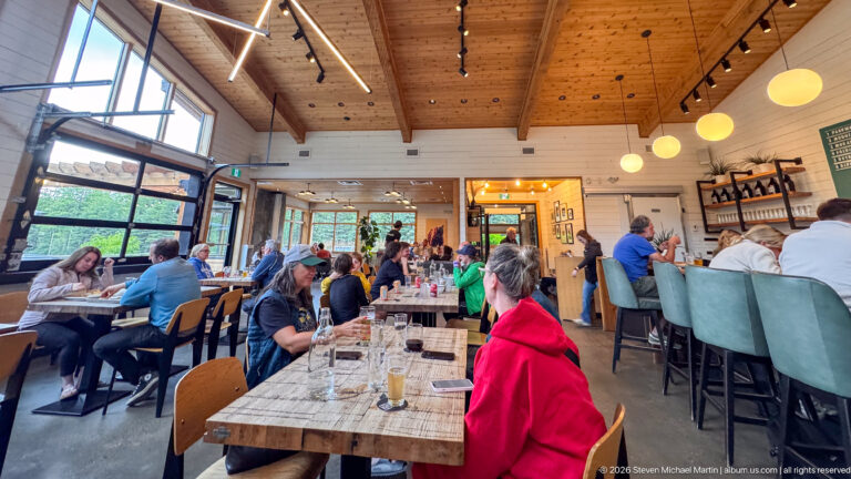 Busy dining room with communal wood tables, bar seating, globe lights, large windows, and a high timber ceiling.