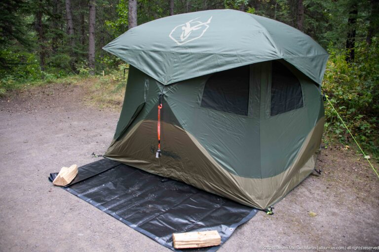 Large green freestanding tent on a black groundsheet at a gravel campsite surrounded by forest.