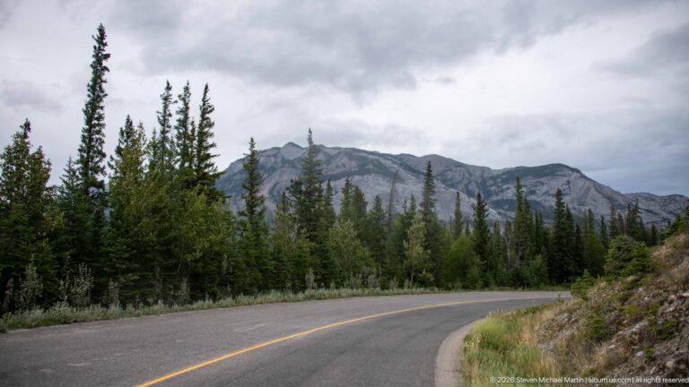 Curving paved mountain road through conifer forest with a long gray rocky ridge rising beyond.