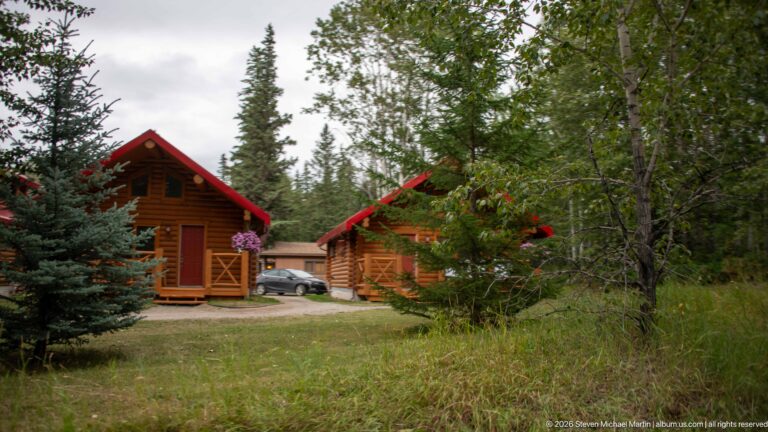 Rustic wood cabins with red gable roofs set among spruce trees, deciduous vegetation, and grass.