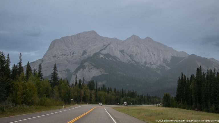 Two-lane highway curving toward a large pale-gray layered mountain ridge beneath an overcast sky.