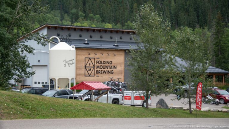 Folding Mountain Brewing building beyond parked vehicles, with its logo and a large white brewing vessel visible.