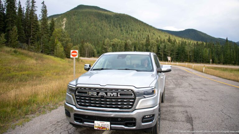 Silver RAM pickup parked beside a paved mountain road with conifer forest and rounded mountain slopes behind.