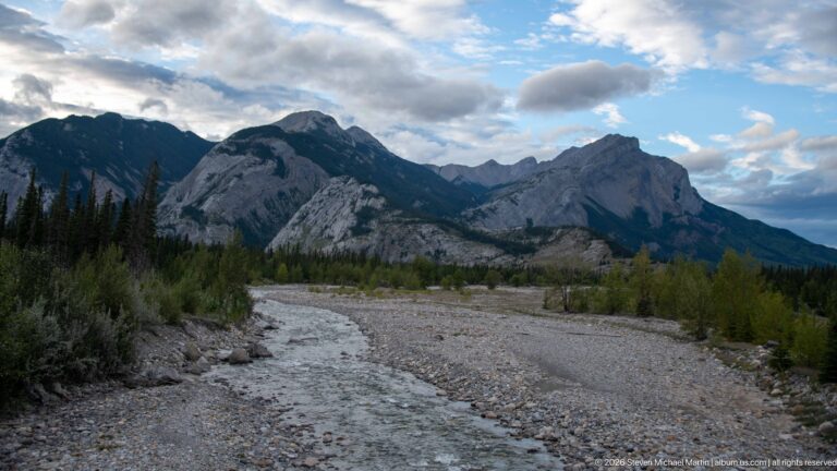 Shallow stream winding through a broad gravel bed beneath rocky, forested mountain slopes.