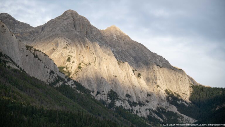 Steep pale-gray layered mountain face lit by sun above dark conifer forest beneath broken clouds.