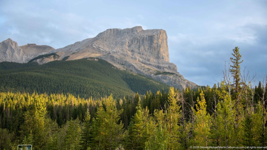 Massive gray layered mountain with steep cliffs rising above dense green conifer forest.