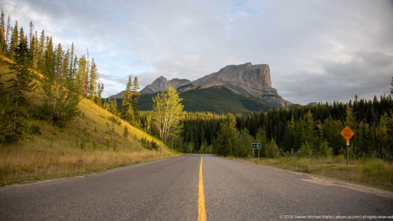 Two-lane mountain road leading through forest toward a massive cliffed peak beneath cloudy evening sky.