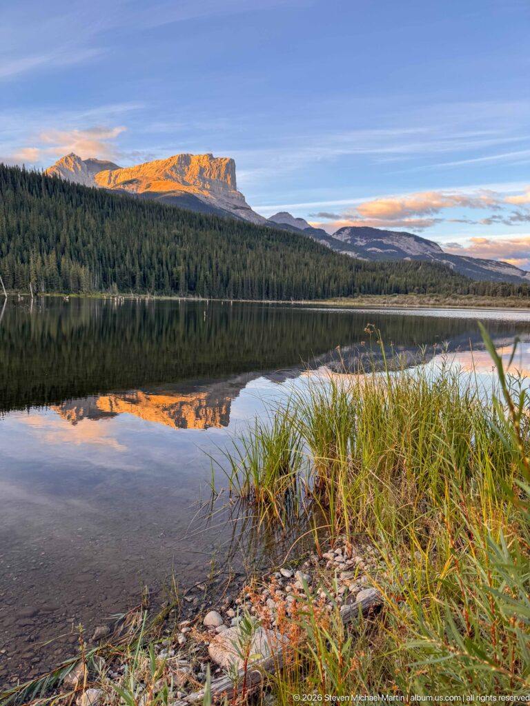 Pebbled shallow shoreline with grasses beside a calm lake reflecting forest and a warm-lit mountain ridge.