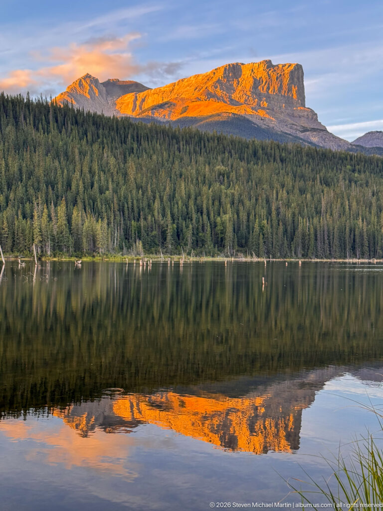 Orange-lit mountain ridge and dark conifer forest mirrored across a calm lake at sunset.