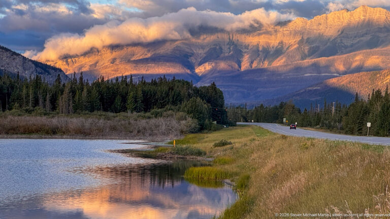 Cloud-wrapped mountain glowing at evening beyond reflective water, forest, grasses, and a paved road.