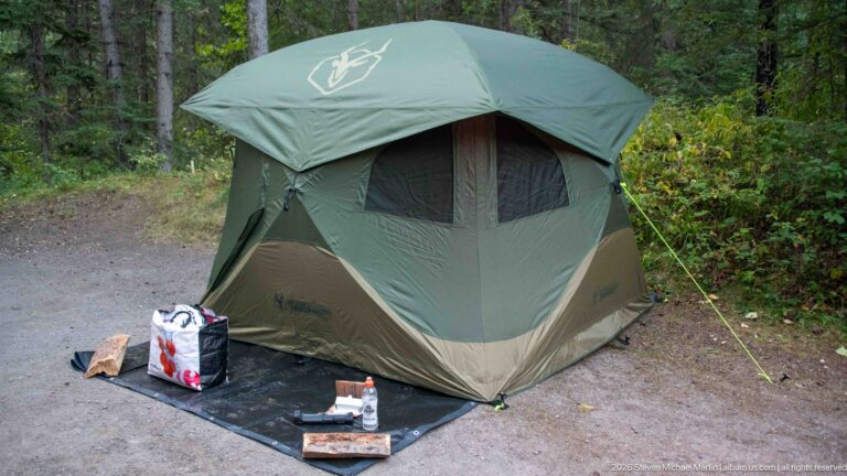 Large green tent with ground mat and supplies pitched on a gravel campsite amid dense forest.