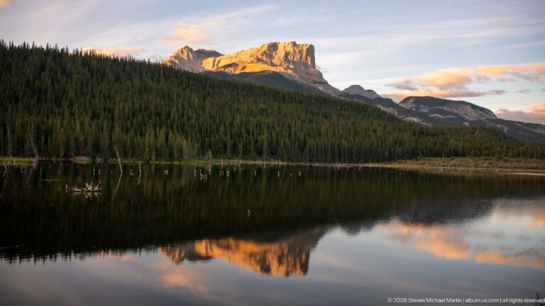 Golden-lit cliffed mountain above dark conifer forest, reflected in a calm lake at evening.