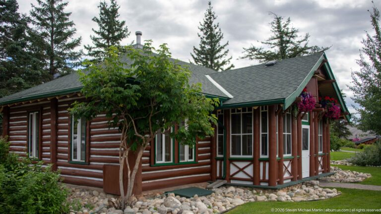 Corner view of brown log cabin with green roof and trim, decorative entrance bay, tree, and stone border.