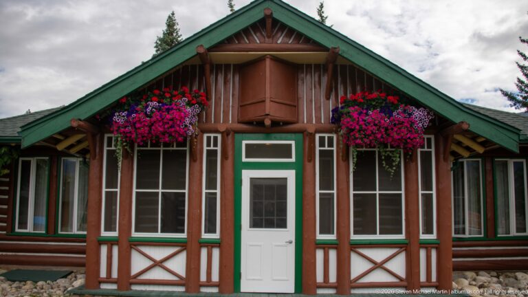 Symmetrical brown-and-green lodge cabin façade with white door, tall windows, and two pink hanging baskets.