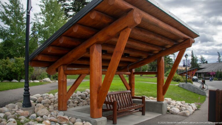Open log shelter with a wooden bench on a concrete pad edged by rounded river stones.