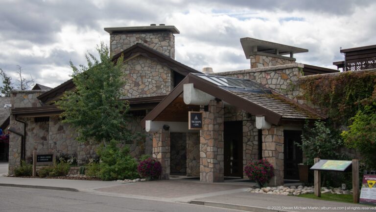 Stone Jasper Park Lodge building with sheltered entrance, broad chimneys, timber roof, trees, and flowers.