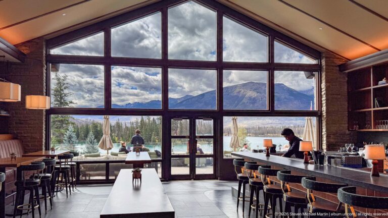 Large lodge windows framing Lac Beauvert and mountains, with tables, bar stools, and terrace visitors.