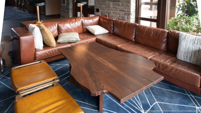 Brown leather sectional surrounding a large live-edge wood table on a dark patterned rug.