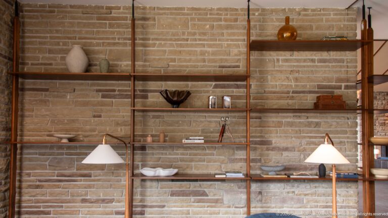 Open metal-and-wood shelves with ceramics and books against a pale stone wall, flanked by lamps.