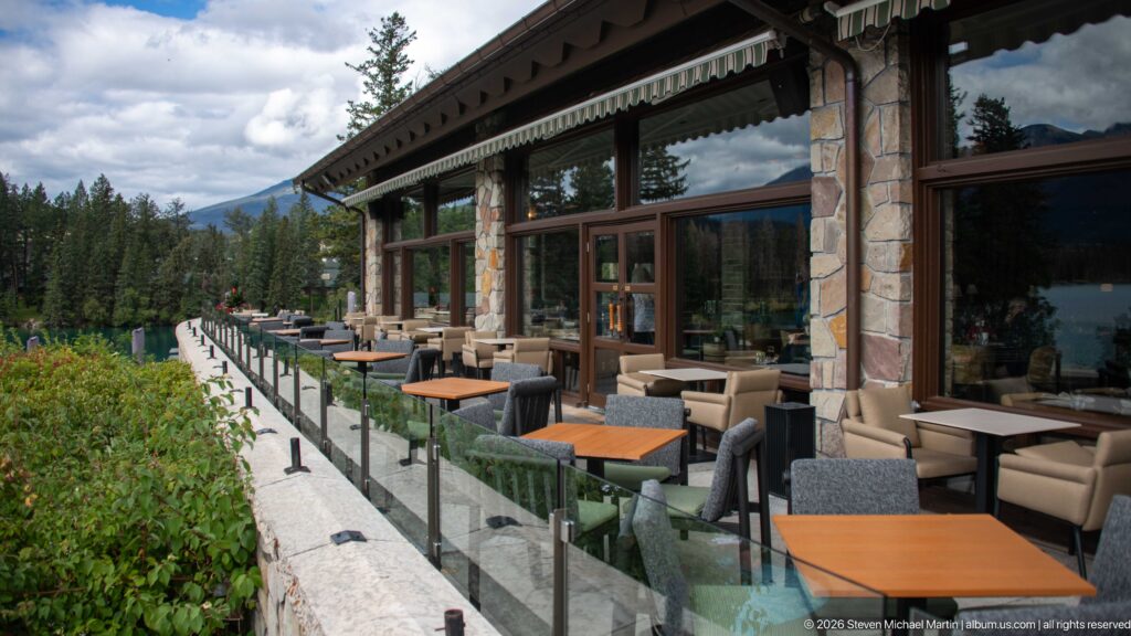 Outdoor lodge terrace with cushioned chairs, small tables, glass railing, stone piers, and large windows.