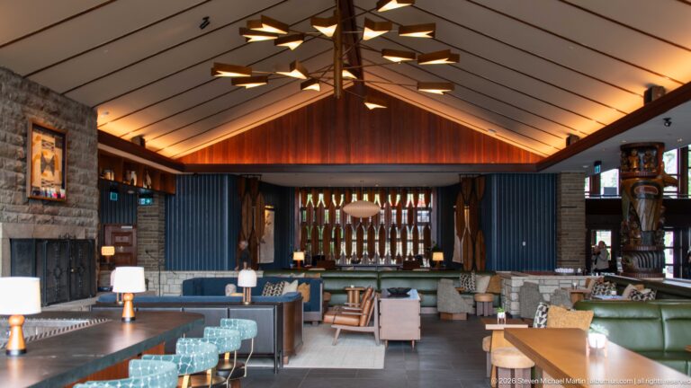 Large lodge lounge with stone fireplace, carved wooden columns, upholstered seating, and geometric ceiling light.