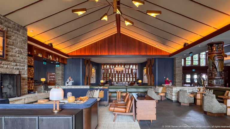 Symmetrical lodge lounge with pitched ceiling, geometric light fixture, stone walls, seating, and carved columns.