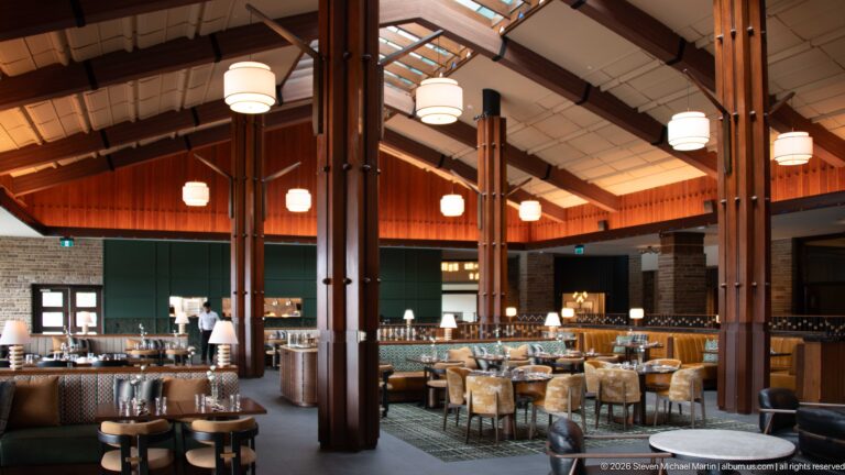 Dining room with massive timber columns, pendant lights, wood paneling, stone walls, and grouped tables.