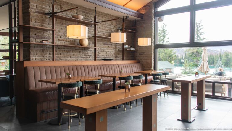 Modern Jasper Park Lodge dining area with wood tables, banquette seating, stone wall, and large lakeside windows.