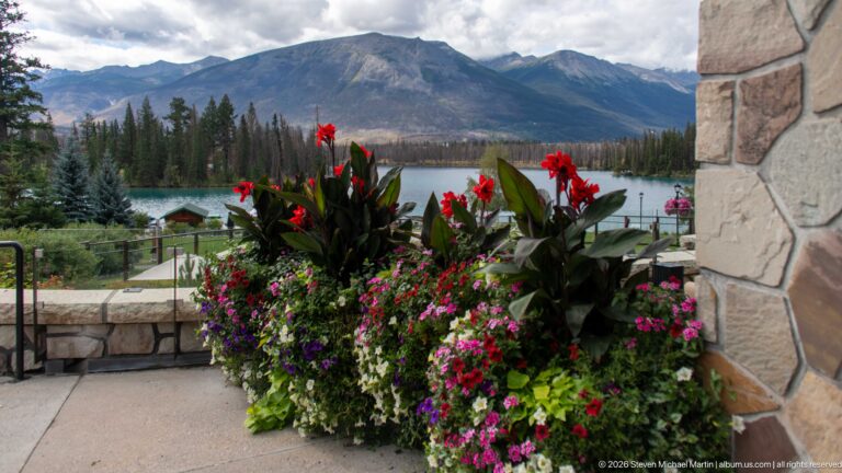 Large red and magenta flower planter on a stone terrace overlooking Lac Beauvert and mountains.