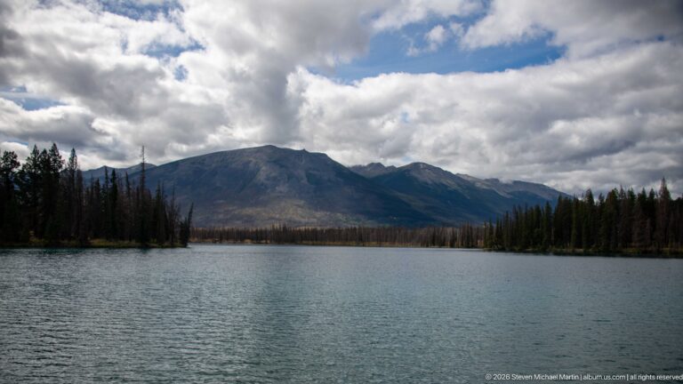 Rippling Lac Beauvert bordered by conifers beneath a broad mountain ridge and broken clouds.