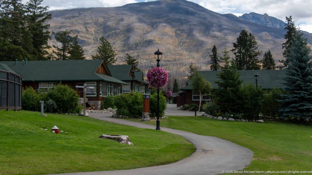 Low green-roofed lodge cabins along a curved path with conifers and a mountain backdrop.