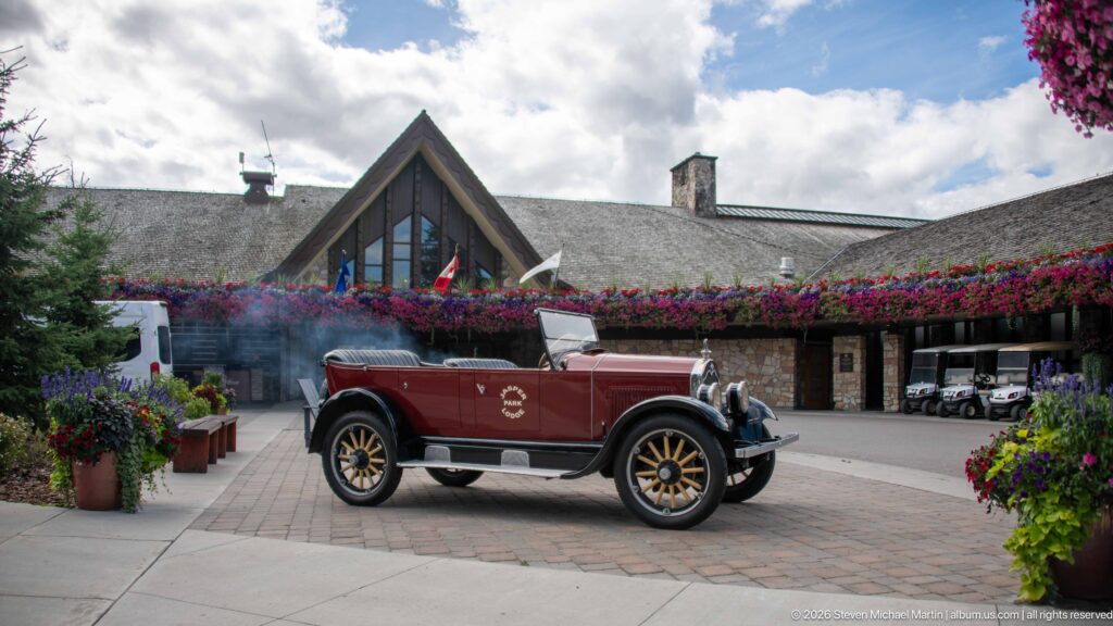Burgundy open touring car centered before Jasper Park Lodge, framed by abundant summer flowers.