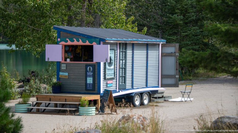 Small blue-gray mobile kiosk with open service window beside a wooded lakeshore.