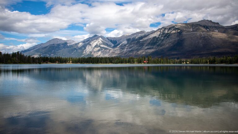 Rugged mountains and clouds reflected in a calm lake beyond a narrow conifer-lined shore.