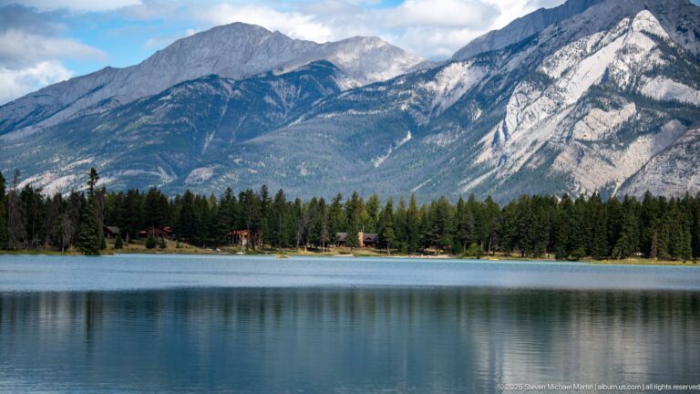 Calm lake with small shoreline buildings beneath steep, striated Rocky Mountain slopes.