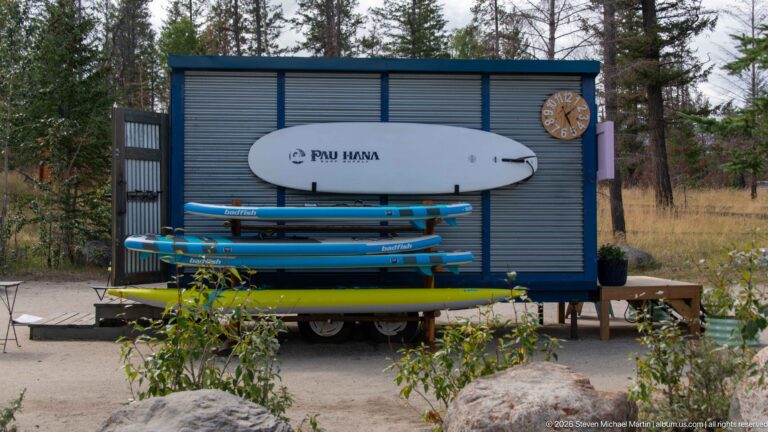Stacked colorful paddleboards on a rack beside a small corrugated lakeside utility structure.