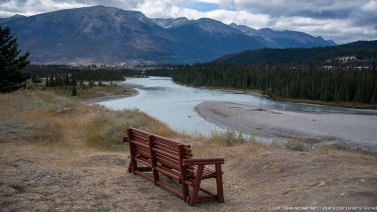 Pair of red chairs overlooking a winding turquoise river beneath dark Rocky Mountain slopes.