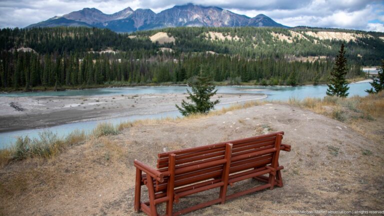 Two red wooden chairs facing a turquoise river, gravel bars, forest, and distant mountains.
