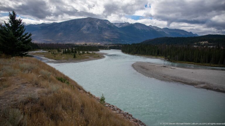 Curving turquoise river and gravel bars leading toward dark mountain peaks beneath cloudy sky.