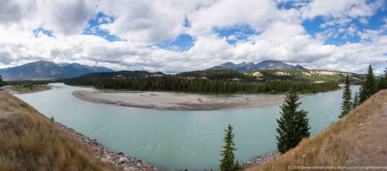 Turquoise mountain river curving around broad gravel bars below forested Rocky Mountain slopes.