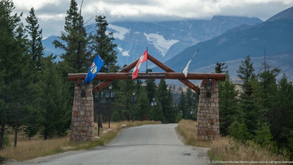 Timber gateway on stone piers spanning a road, with Alberta and Canadian flags and mountains beyond.