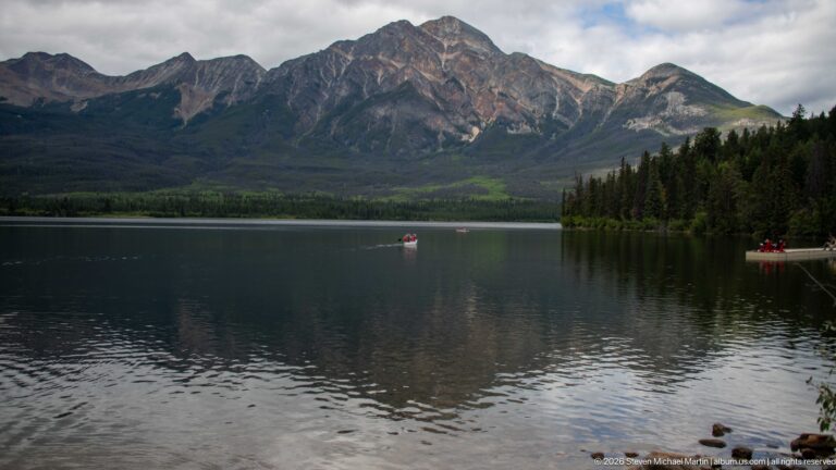 Calm Pyramid Lake reflecting a rugged mountain, with small boats and a forested point at right.