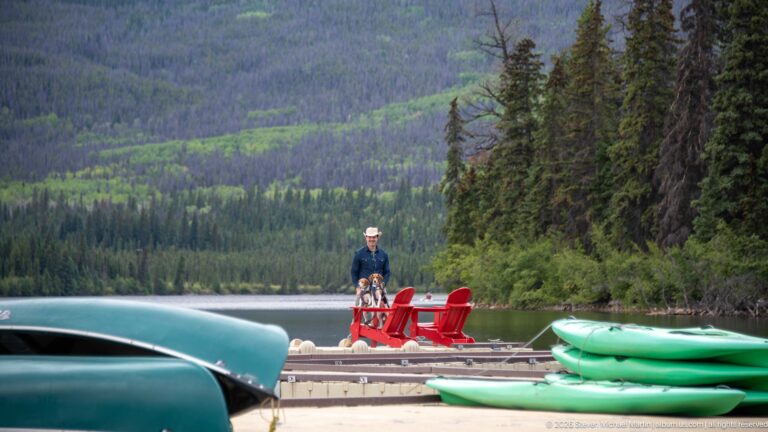 Canoes and stacked green kayaks frame a floating dock with red chairs and a visitor beyond.