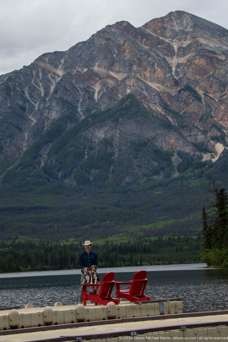 Small figures and red chairs on a lake dock below a massive rocky mountain slope.