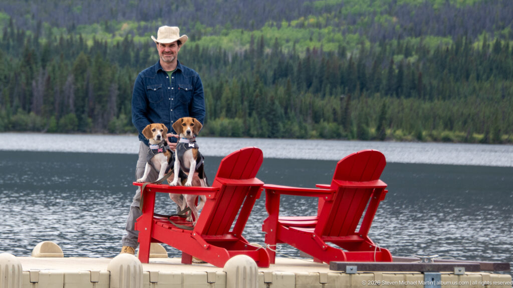 Man standing behind two beagles on bright red Adirondack chairs on a floating lake dock.