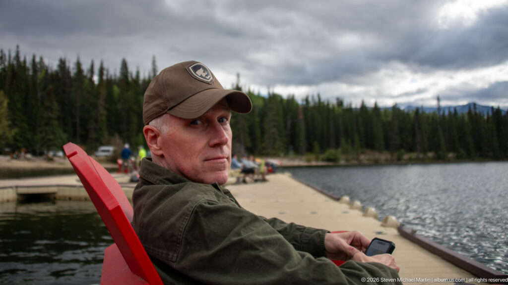 Man seated in a red Adirondack chair on a dock beside Pyramid Lake, with forest across the water.