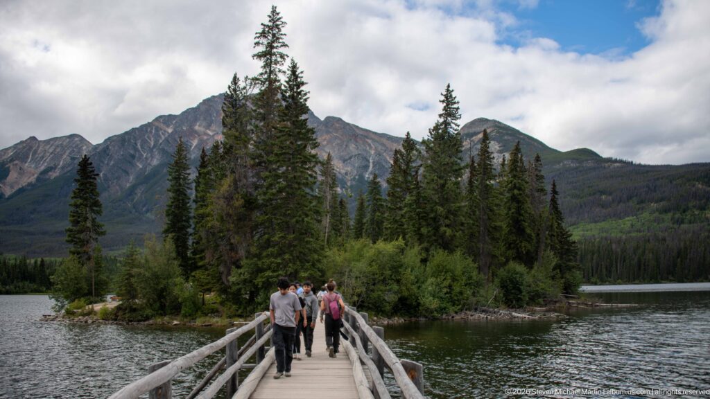 People walk away along a wooden bridge toward conifers on Pyramid Island, backed by rugged mountains.
