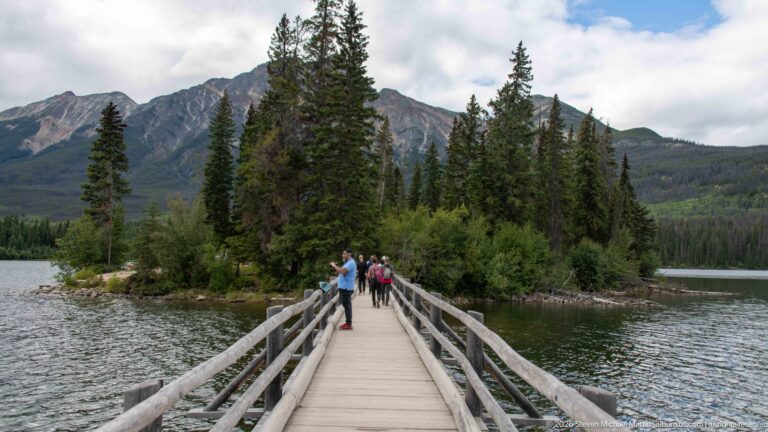 Visitors walk along a wooden footbridge toward a forested island with mountains rising behind.
